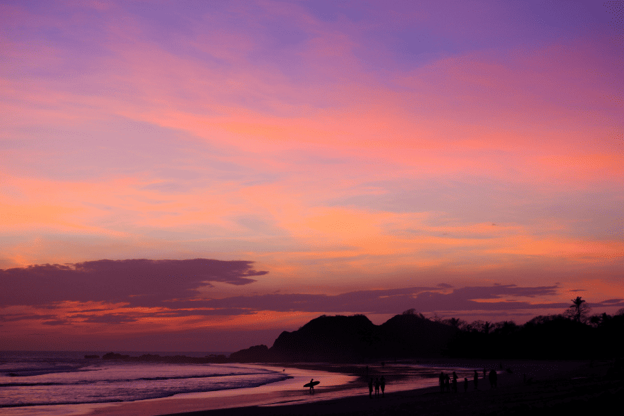 An image with a photo of a coastal sunset in Nosara, Costa Rica. The sky is in shades of purple and orange, and trees and people along the beach are silhouetted in black.